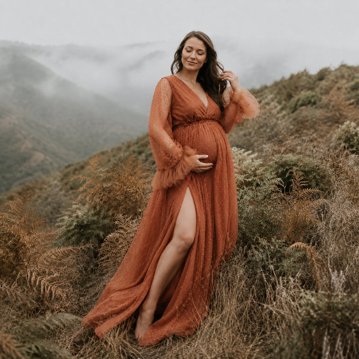 A pregnant woman stands on a misty, fern-covered hillside wearing a Alectrona terracotta polka dot maternity gown with sheer long sleeves, ruffled cuffs, a V neckline, and a high side slit that reveals her leg against the natural backdrop.