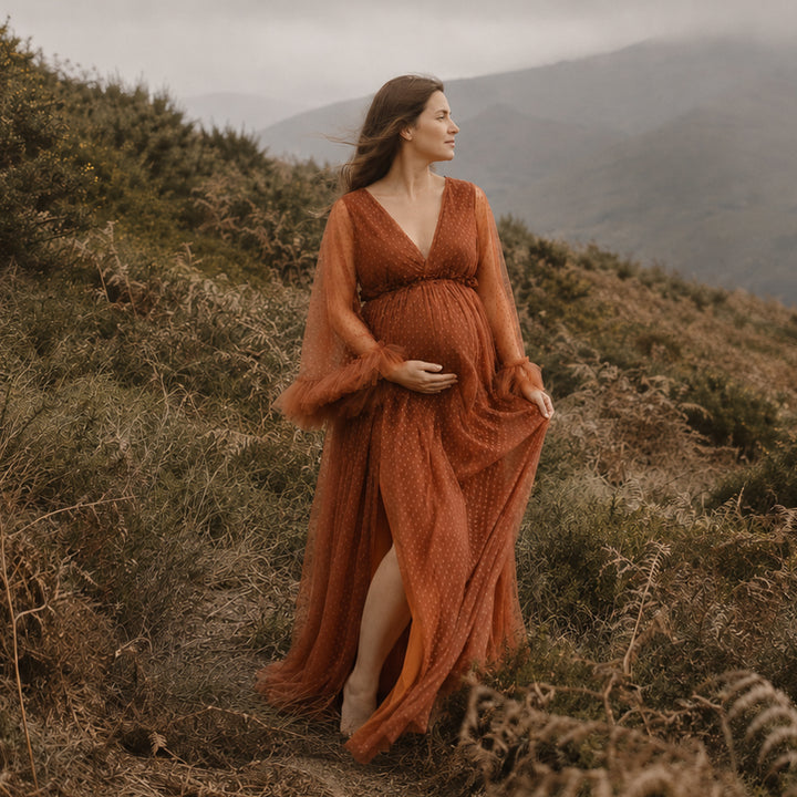 A pregnant woman stands on a misty, wearing a Alectrona terracotta polka dot maternity gown with sheer long sleeves,ruffled cuffs, a V neckline.