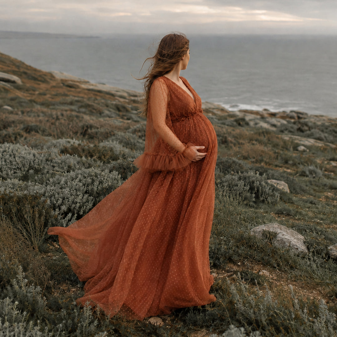 A pregnant woman stands in profile on a rocky coastal cliff wearing a Alectrona terracotta polka dot tulle maternity gown with sheer long sleeves, ruffled cuffs, and a V-neckline.