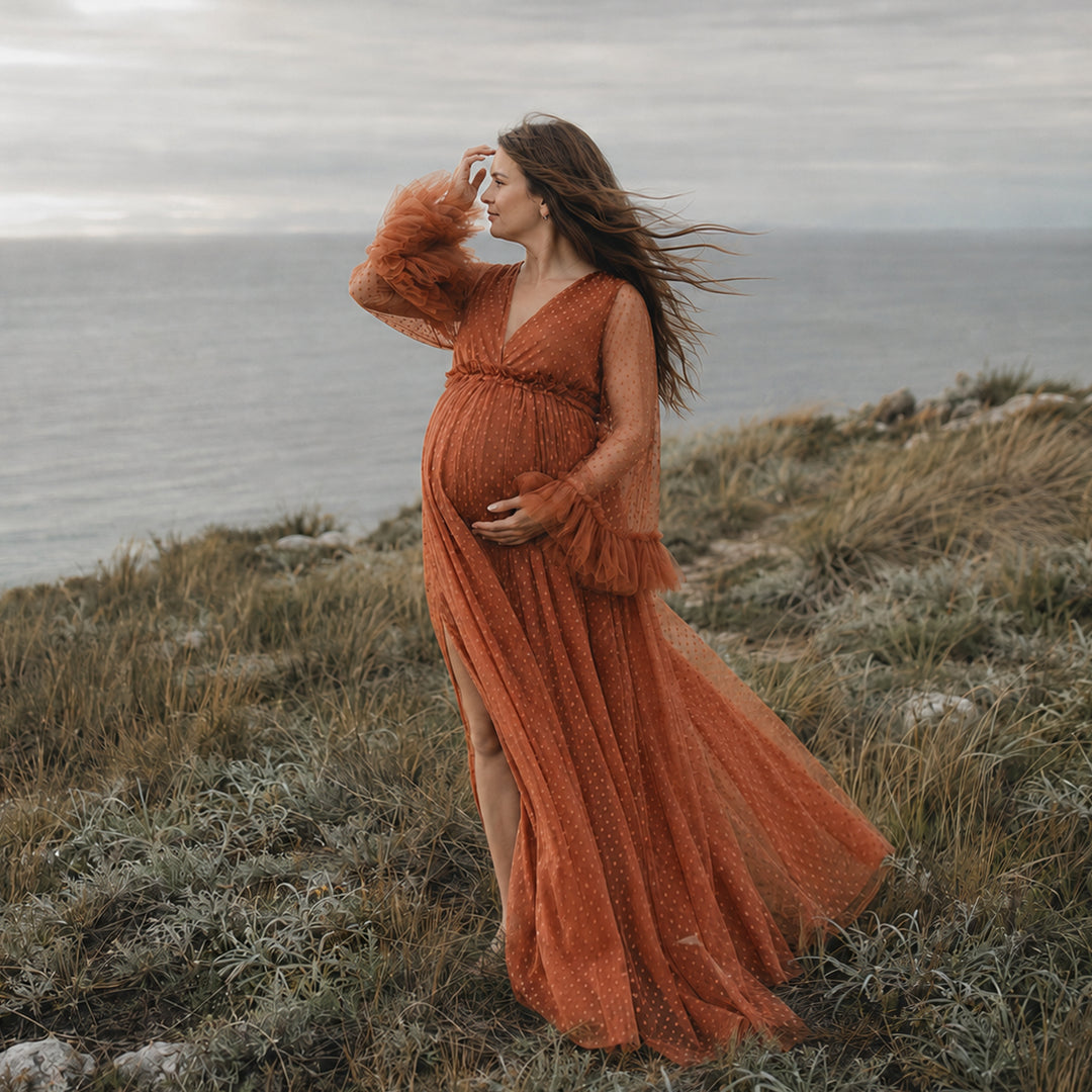 A pregnant woman stands in profile on a rocky coastal cliff wearing a Alectrona terracotta polka dot  tulle maternity gown with sheer long sleeves, ruffled cuffs, and a V-neckline.