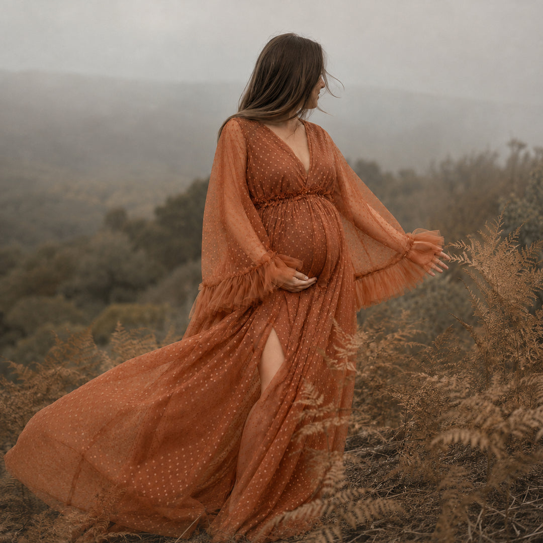 A pregnant woman stands on a hillside wearing a Alectrona terracotta polka dot  tulle maternity gown with long ruffled sleeves, a V-neckline, and a high side slit.