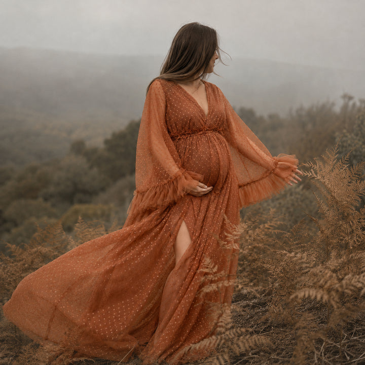 A pregnant woman stands on a hillside wearing a Alectrona terracotta polka dot  tulle maternity gown with long ruffled sleeves, a V-neckline, and a high side slit.