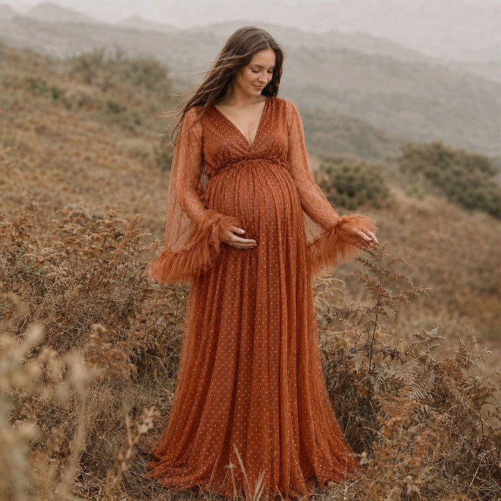 A pregnant woman stands in a grassy landscape wearing a Alectrona terracotta polka dot  tulle maternity gown with long ruffled sleeves, a V-neckline, and a full skirt that spreads across the ground.