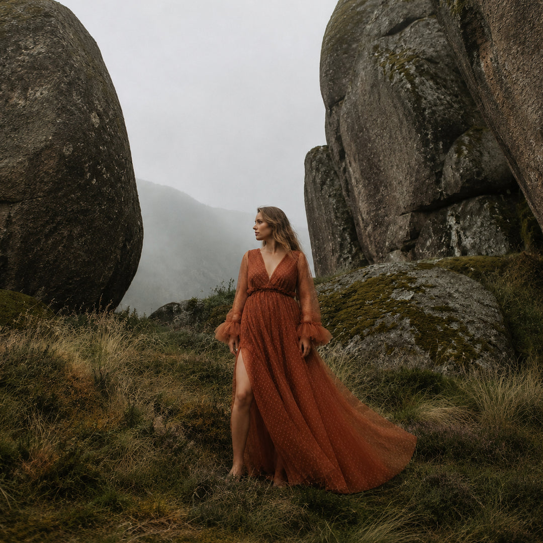 A pregnant woman stands between large boulders on a grassy hillside wearing a Alectrona terracotta polka dot tulle maternity gown with long ruffled sleeves, a V-neckline, and a high side slit.