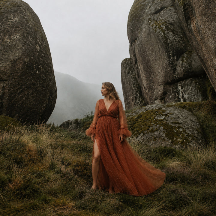A pregnant woman stands between large boulders on a grassy hillside wearing a Alectrona terracotta polka dot tulle maternity gown with long ruffled sleeves, a V-neckline, and a high side slit.