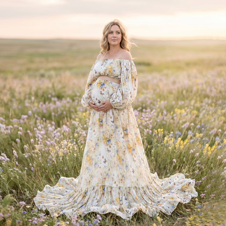 A pregnant woman in an Aurelia floral and butterfly print off-shoulder two-piece stands in a flower field holding her belly.
