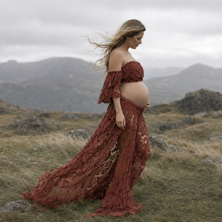 A pregnant woman stands in profile on a windswept grassy hill wearing a Bendis terracotta two-piece lace maternity set with off-the-shoulder ruffled sleeves and a high-slit skirt that reveals her bare baby bump against a mountain backdrop.