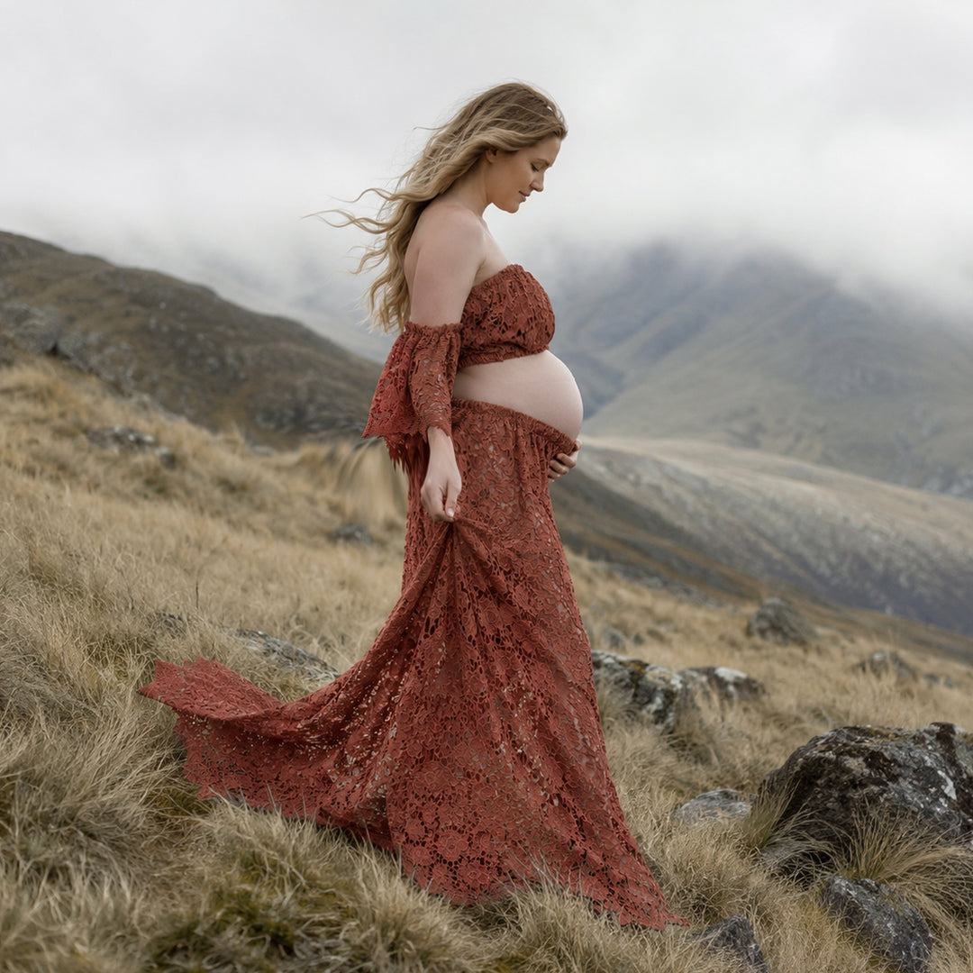 A pregnant woman stands in profile on a misty, grassy hillside wearing a Bendis terracotta two-piece lace maternity set with off-the-shoulder ruffled sleeves and a high-waisted skirt that reveals her bare baby bump against a mountain backdrop.