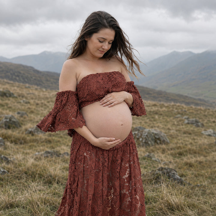 A pregnant woman stands in profile on a misty, grassy hillside wearing a Bendis terracotta two-piece lace maternity set with off-the-shoulder ruffled sleeves and a high-waisted skirt that reveals her bare baby bump against a mountain backdrop.