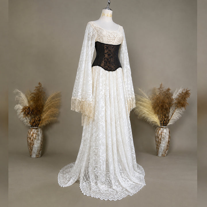 A long beige lace dress displayed on a mannequin against a warm taupe studio background, with a black lace corset bodice and two long sheer lace sleeves with fringe. Two vases with dried pampas grass are placed on either side.