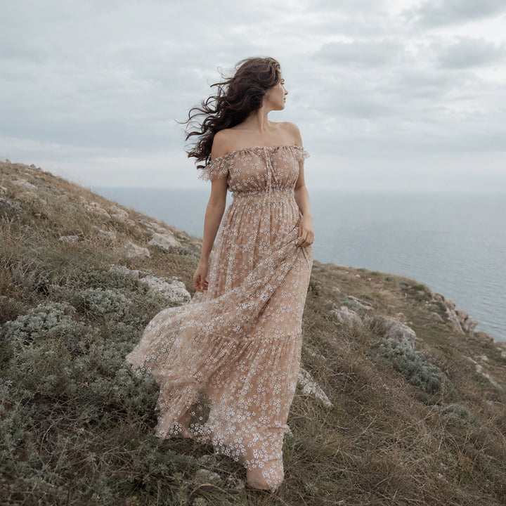 A woman stands on a grassy coastal cliff wearing a Clete khaki off-shoulder tulle maxi dress with white floral embroidery, ruffled neckline, and flowing skirt.