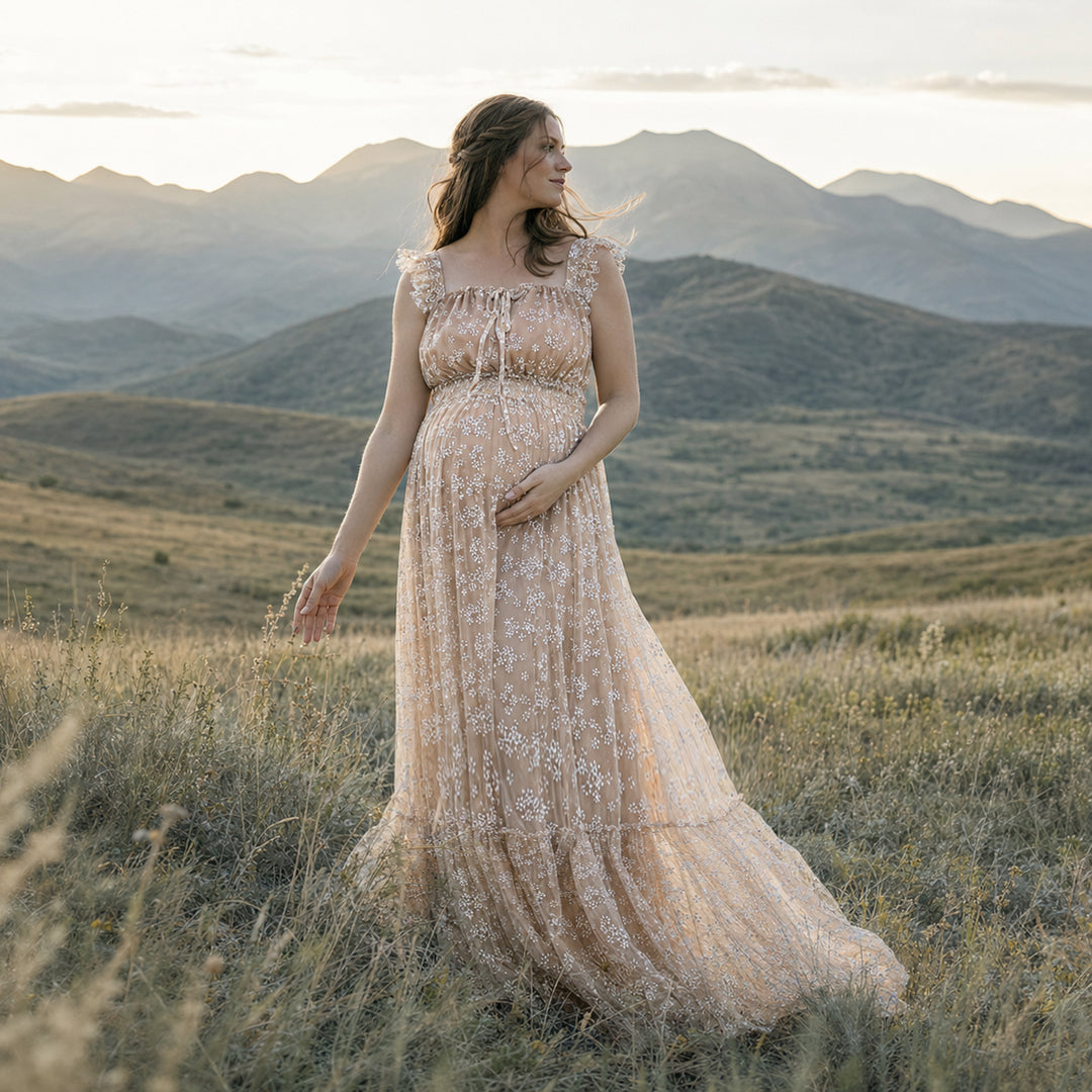 A pregnant woman stands in a grassy mountain landscape wearing a Clete khaki floral embroidered tulle maternity gown with ruffled cap sleeves and a flowing skirt.