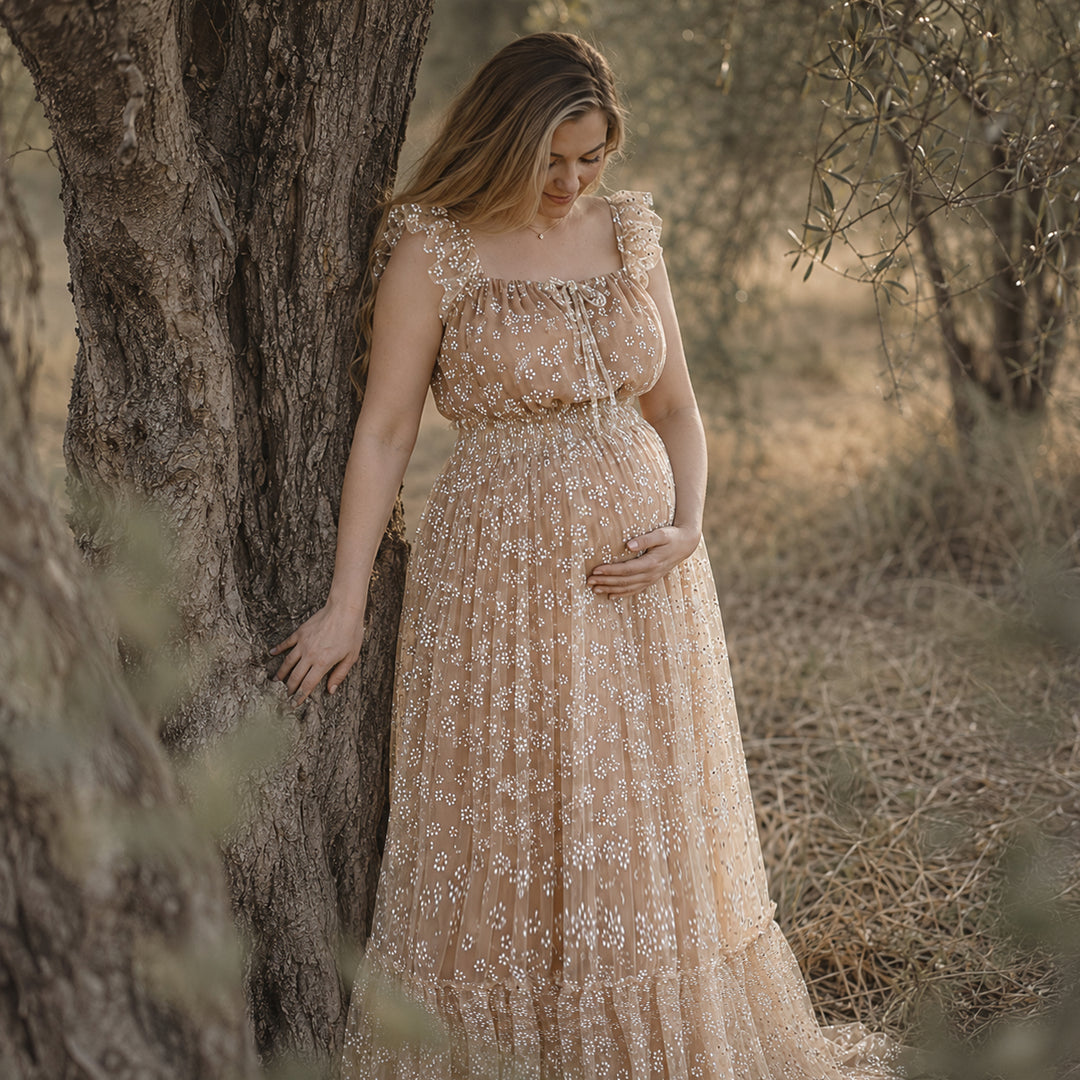 A pregnant woman leans against a tree trunk in a sunlit grove, wearing a Clete khaki floral embroidered tulle maternity gown with ruffled square neckline and tiered skirt.