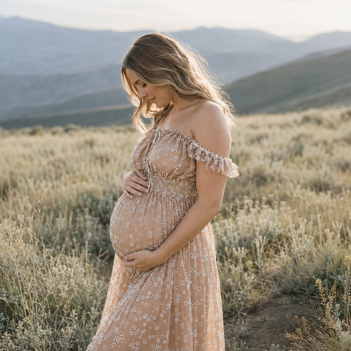 A pregnant woman stands in a grassy mountain landscape wearing a Clete khaki floral embroidered tulle maternity gown with ruffled cap sleeves and a flowing skirt.