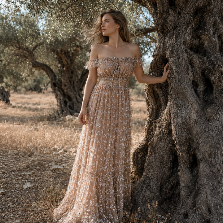 A woman stands beside a gnarled olive tree in a sun-dappled grove, wearing a Clete sheer khaki off-the-shoulder tulle gown with delicate white floral embroidery and ruffled short sleeves that flows to the rocky ground.