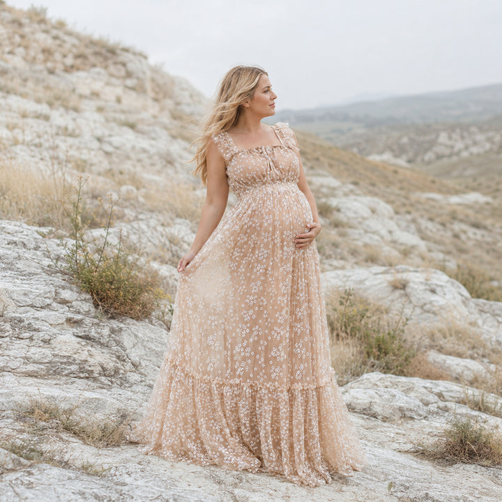 A pregnant woman stands on a rocky outcrop in a rugged, hilly landscape wearing a Clete flowing beige floral embroidered tulle maternity gown with ruffled cap sleeves and a tiered skirt that drapes over the stones.