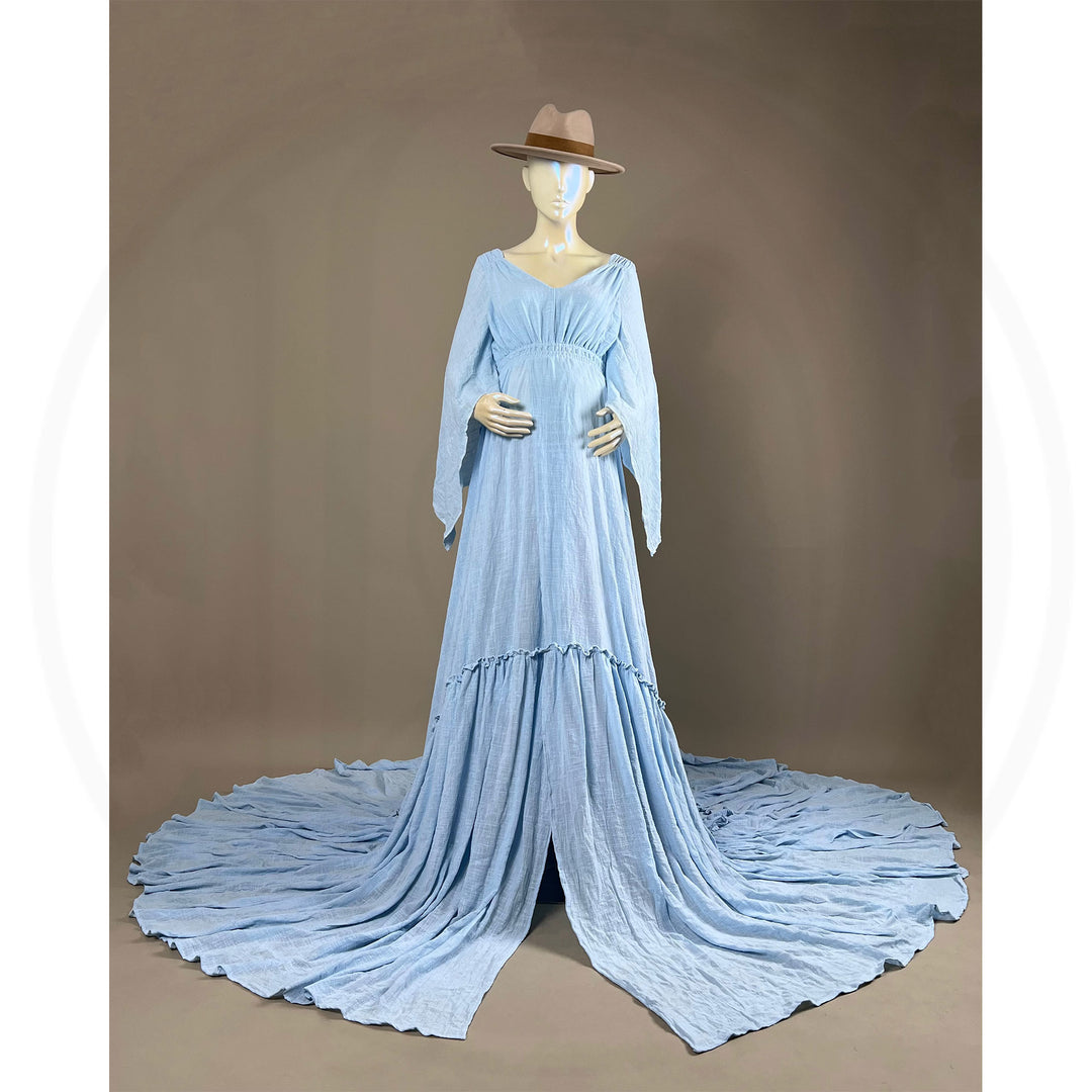 A Daphne light blue long-sleeved V-neck gown is displayed on a mannequin, with hands positioned on the simulated belly. The dress features a high front slit and its hem and train are spread out on the floor.