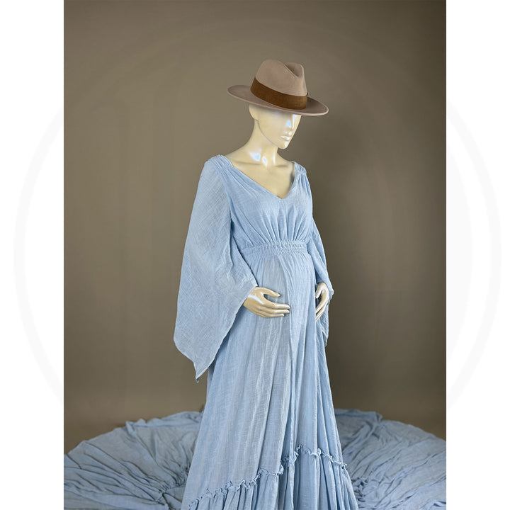 A  Daphne light blue long-sleeved V-neck gown is displayed on a mannequin, with hands positioned on the simulated belly. The dress features a high front slit and its hem and train are spread out on the floor.
