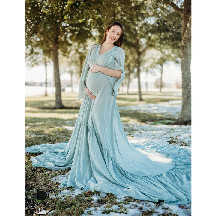 A pregnant woman in a Daphne light blue dress with a shawl stands under trees on snowy grass, hands on her belly.