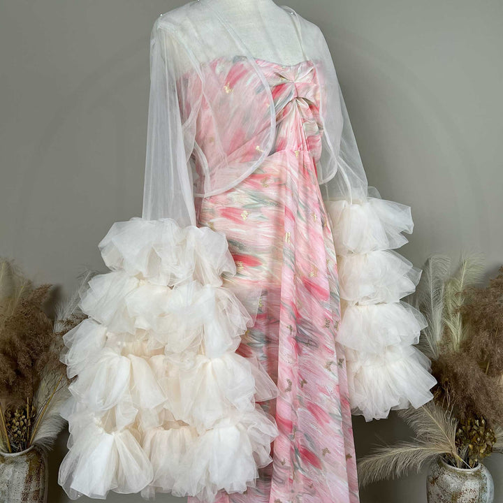 A mannequin displays a side view of a Eileithyia pink printed strapless dress with a light apricot sheer bolero and tiered sleeves; the dress features pleats.