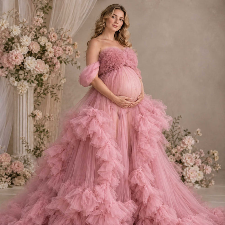 A pregnant woman stands in a Electra pink strapless ruffled tulle gown, holding her belly; the dress features multi-layered ruffles.