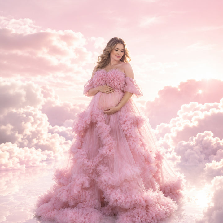 A pregnant woman stands in a Electra pink off-shoulder ruffled gown, holding her belly; the dress features multi-layered ruffles.