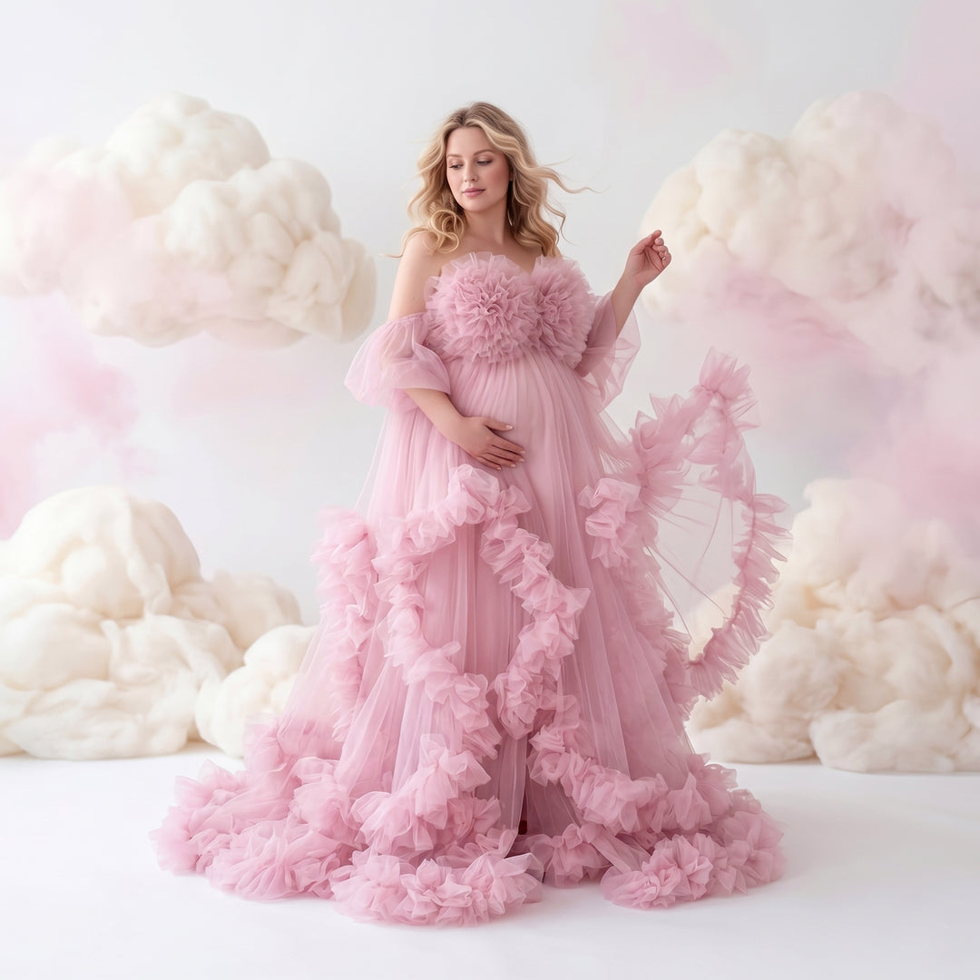 A pregnant woman in a Electra pink off-shoulder ruffled gown holds her belly and lifts the skirt; the dress features multi-layered ruffles.