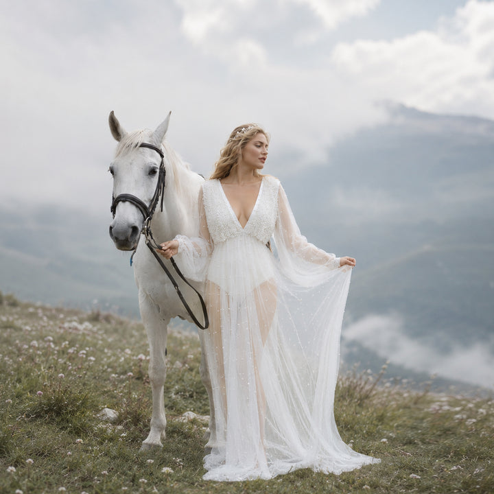 A woman in a Eos ivory deep-V beaded bodysuit with a sheer skirt holds a white horse's reins and lifts the hem.