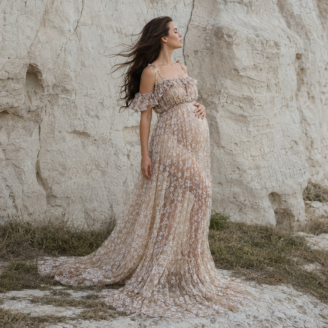 A pregnant woman stands before a Ersa khaki rock face wearing a sheer, floral embroidered off-shoulder gown.