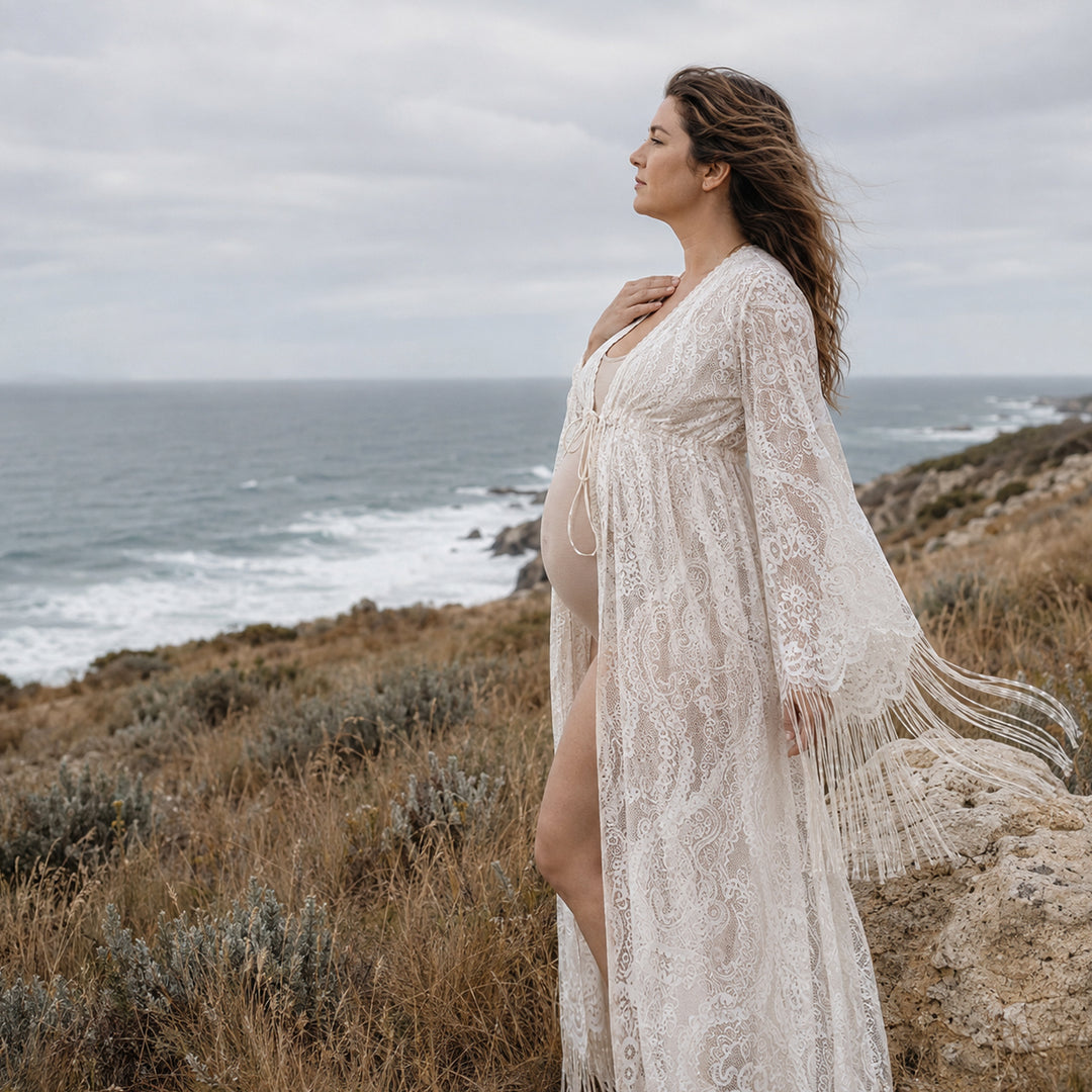 A pregnant woman stands on a grassy hillside near the ocean coast, wearing a Fama beige lace open-front robe with bell sleeves and fringe trim.