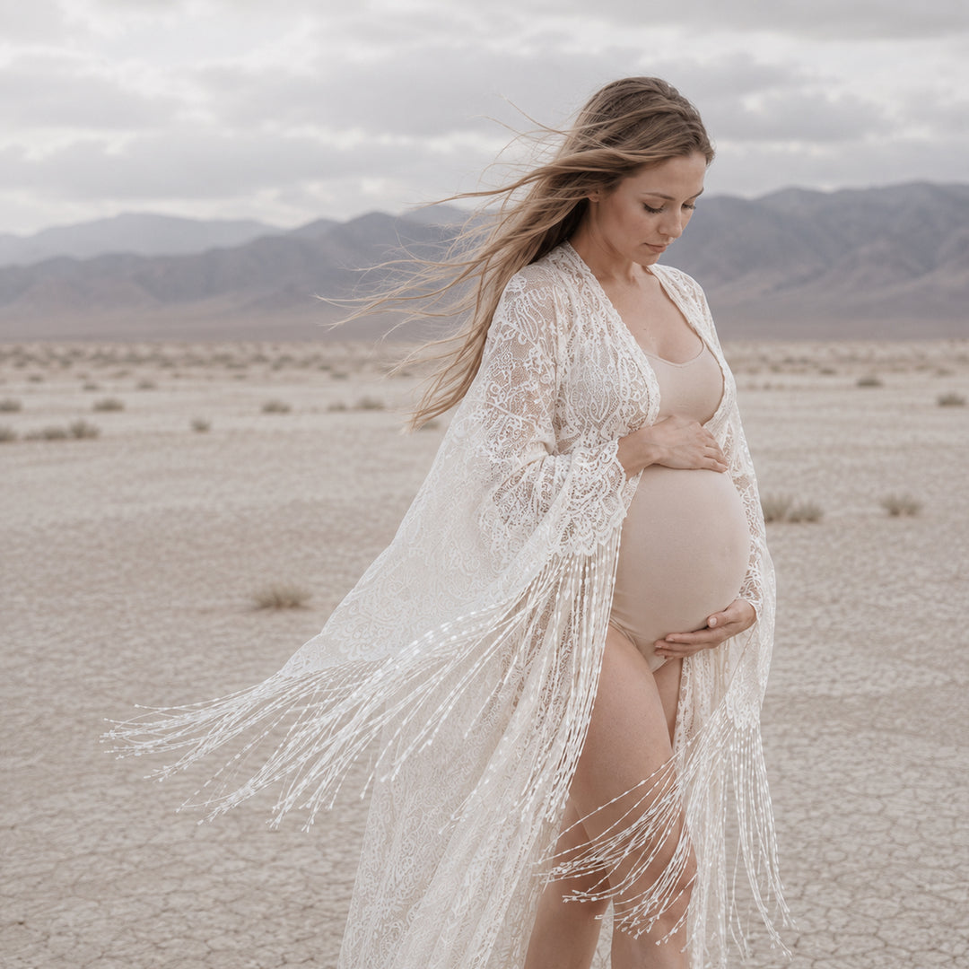 A pregnant woman stands in a dry, cracked desert landscape with mountains in the distance, wearing a Fama beige lace fringe robe over a maternity bodysuit.