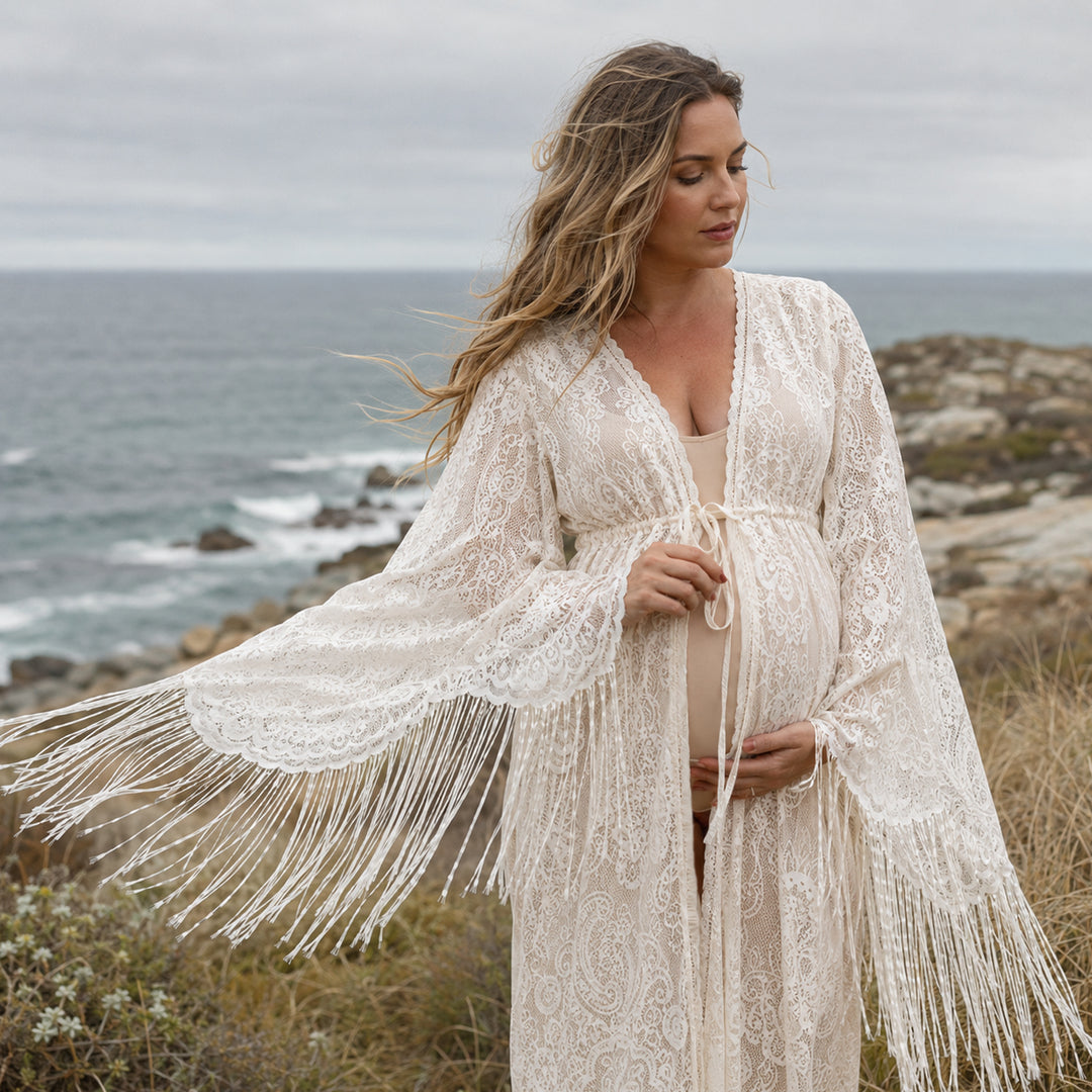 A pregnant woman stands on a grassy coastal cliff overlooking the ocean, wearing a Fama beige lace maternity with long bell sleeves and fringe trim. 