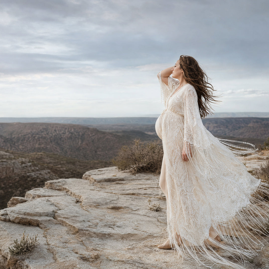 A pregnant woman stands in profile on a rocky cliff edge with a vast canyon landscape in the background, wearing a Fama floor-length beige lace robe with fringe details.