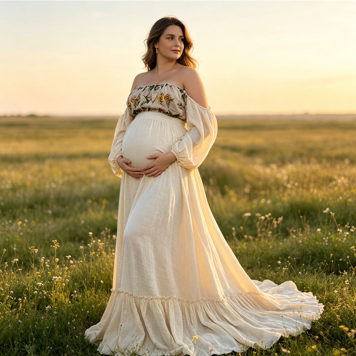 A pregnant woman stands in a grassy field wearing a Freda beige boho embroidery floral two-piece gown, holding her belly.The top is printed, the skirt hem is ruffled, and the background is a vast natural landscape.