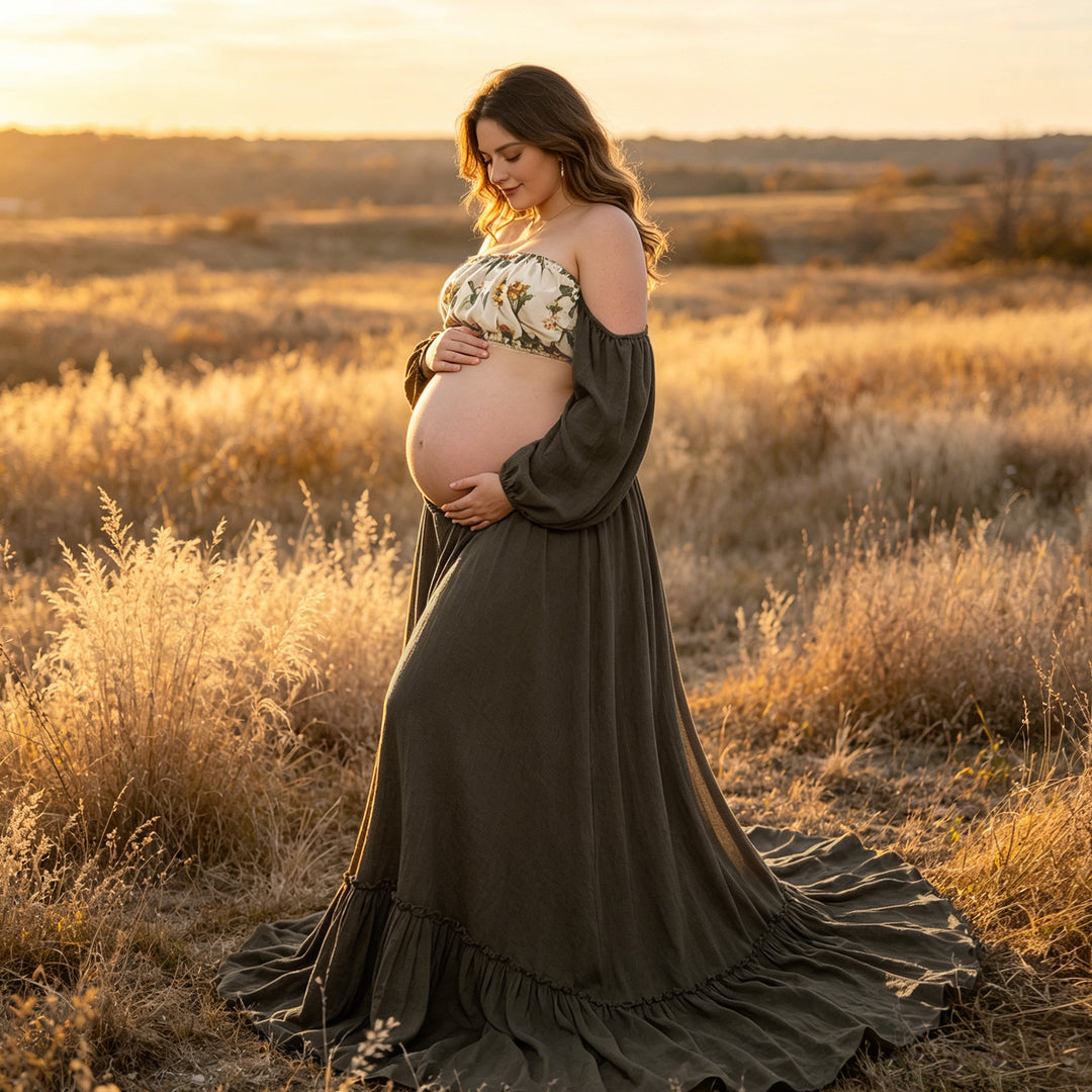 A pregnant woman in a Freda olive gown and floral top stands in tall grass, holding her belly.