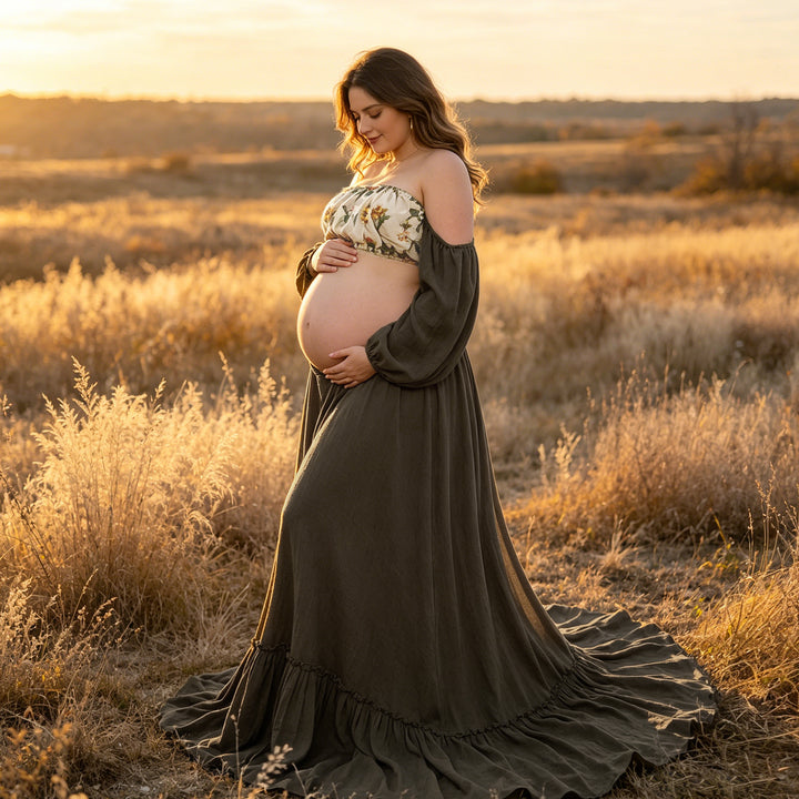 A pregnant woman in a Freda olive gown and floral top stands in tall grass, holding her belly.