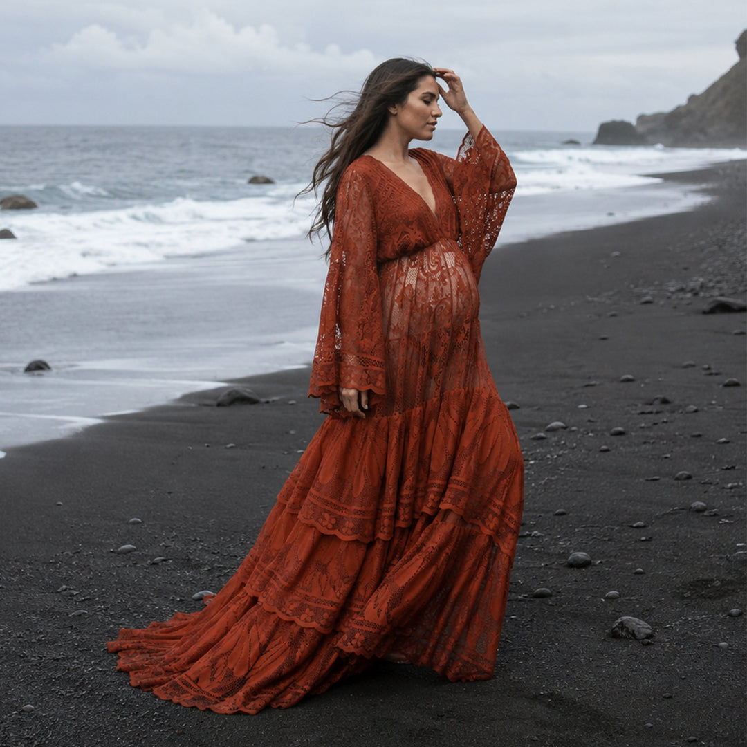 A pregnant woman stands in profile on a rocky black beach near the water's edge, wearing a Harpocrates terracotta lace dress with long sleeves. 