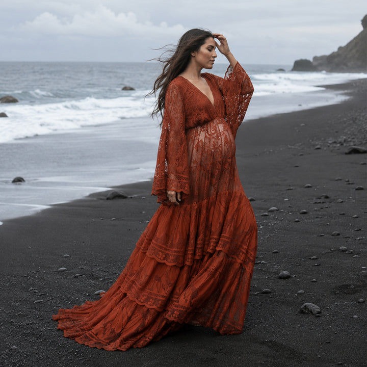 A pregnant woman stands in profile on a rocky black beach near the water's edge, wearing a Harpocrates terracotta lace dress with long sleeves. 