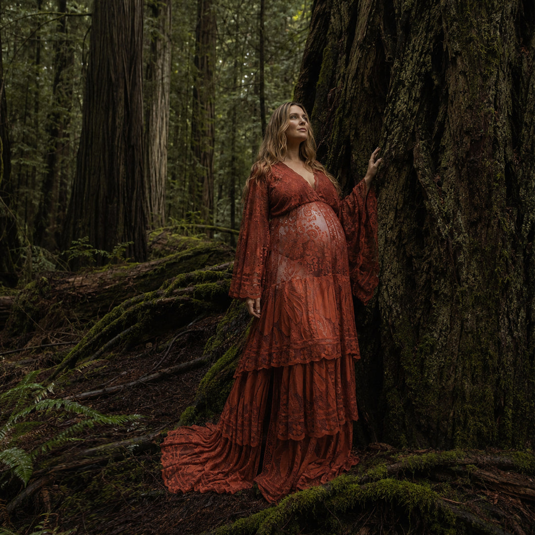 A pregnant woman stands in a dense forest leaning against a large tree trunk covered in moss, wearing a Harpocrates floor length terracotta lace gown.