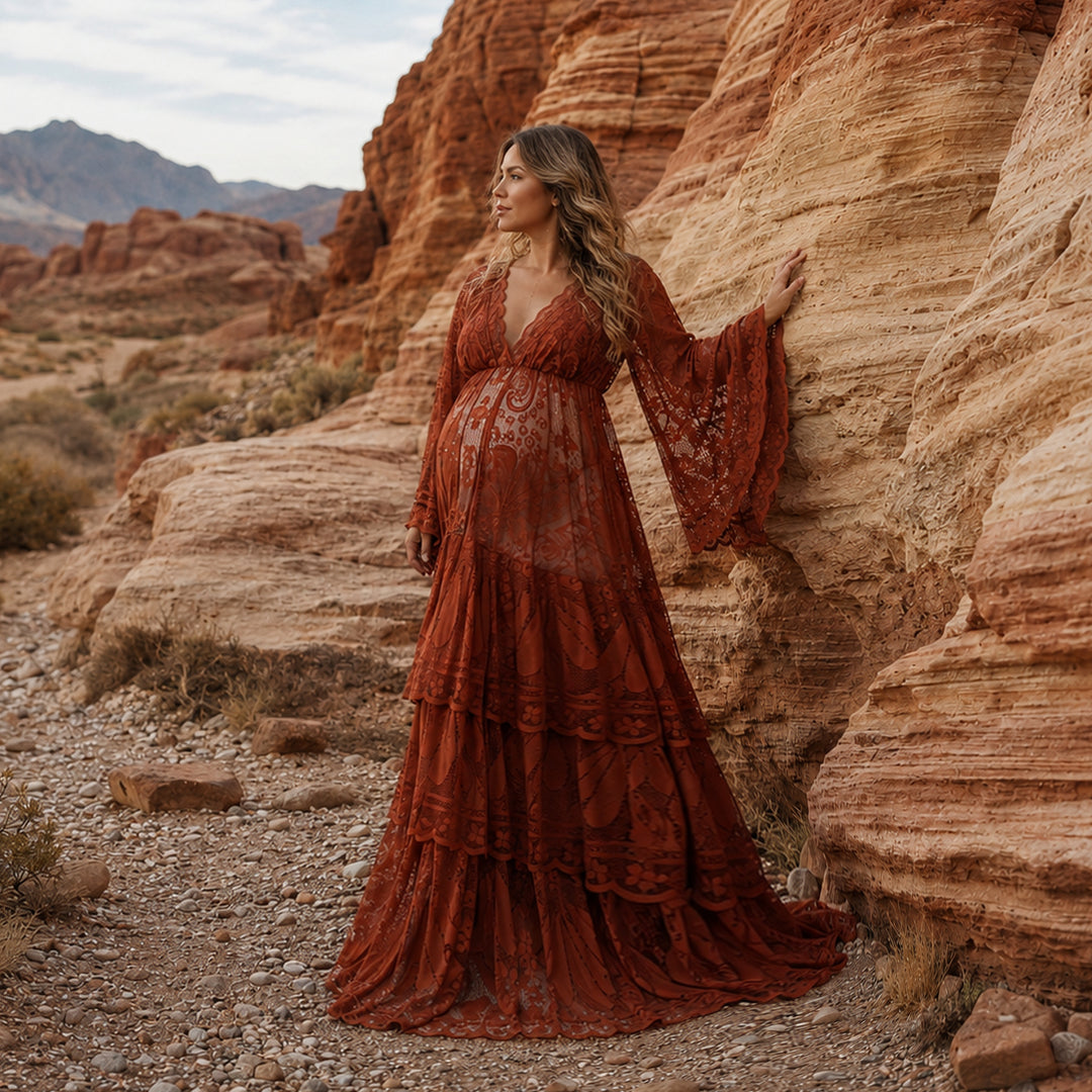 A pregnant woman stands on a gravel path next to layered red rock formations in a canyon landscape, wearing a Harpocrates terracotta tiered lace maternity dress. 