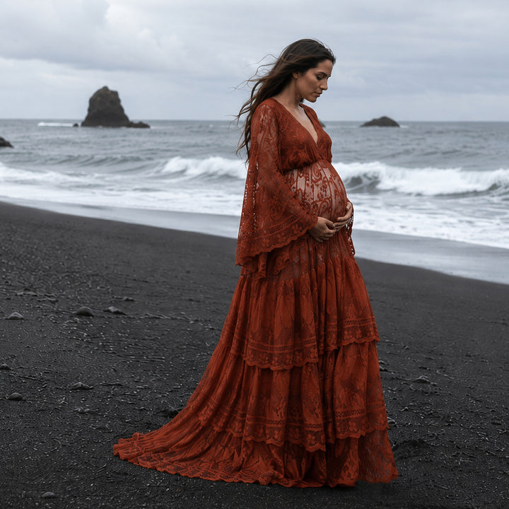 A pregnant woman stands on a black sand beach facing the ocean waves, wearing a Harpocrates terracotta lace maternity gown with a deep V-neckline and tiered skirt. 