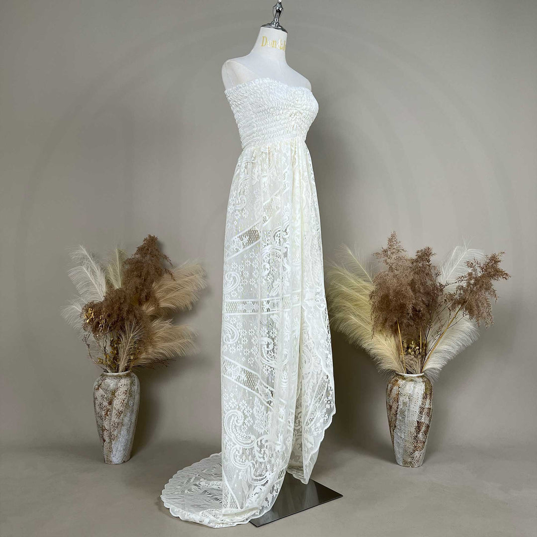 A mannequin shows a side view of a Hippolyta beige strapless lace gown with a ruched bodice and train; the dress features an openwork pattern, set against a gray wall with dried flower vases on both sides.