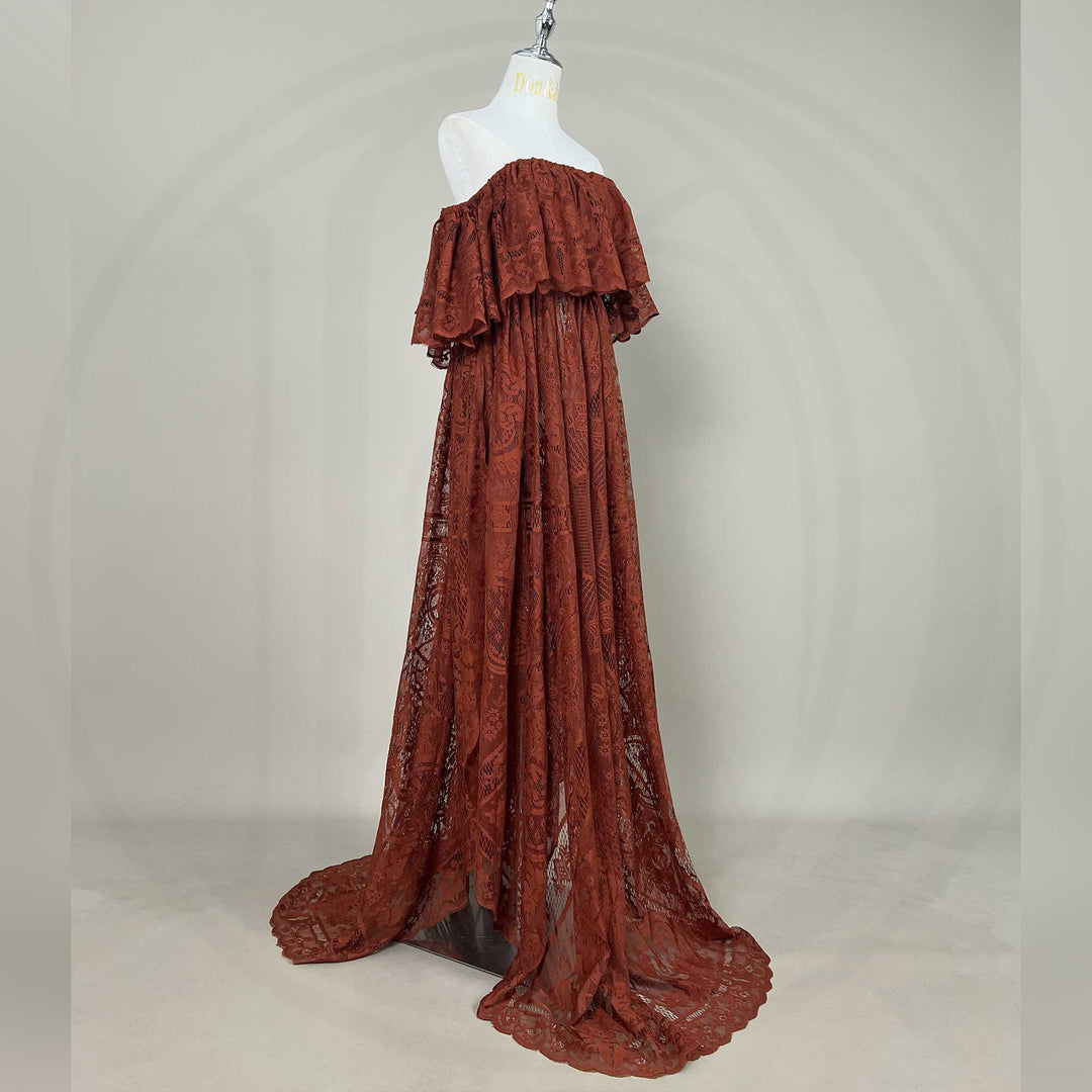 A mannequin a side view of a Kymopoleia terracotta off-shoulder lace gown with ruffled sleeves and a train; the dress features a sheer openwork structure, set against a gray wall with dried flower vases on both sides.