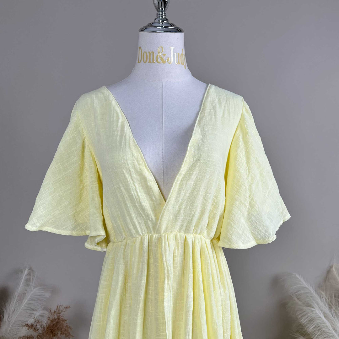 A mannequin displays the front of a Megaera yellow V-neck short-sleeve dress, showing a deep-V cut and waist pleats; the fabric has a textured finish.