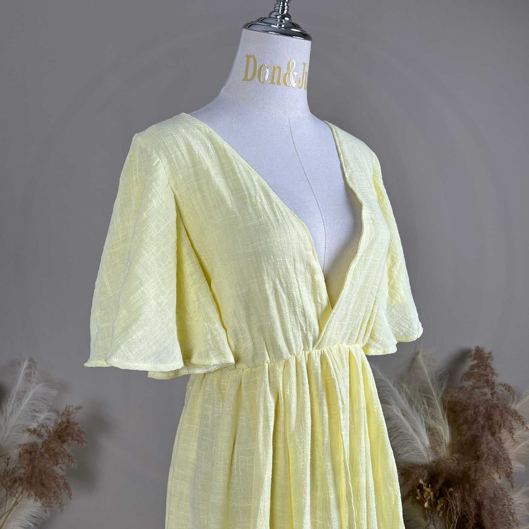 A mannequin displays displays the upper body of yellow V-neck short-sleeve dress, showing a Megaera deep-V cut and waist pleats; the fabric has a textured finish, set against a solid wall with dried flowers on both sides.