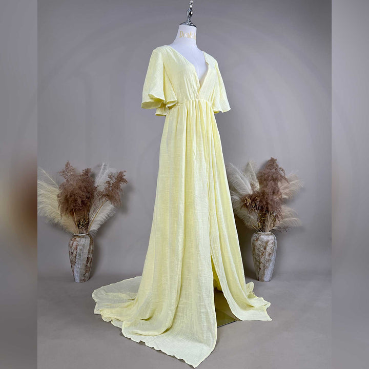 A mannequin shows a side view of a Megaera yellow V-neck short-sleeve gown with a high-waist pleat and train; the fabric has a textured finish, set against a solid wall with dried flower vases on both sides.