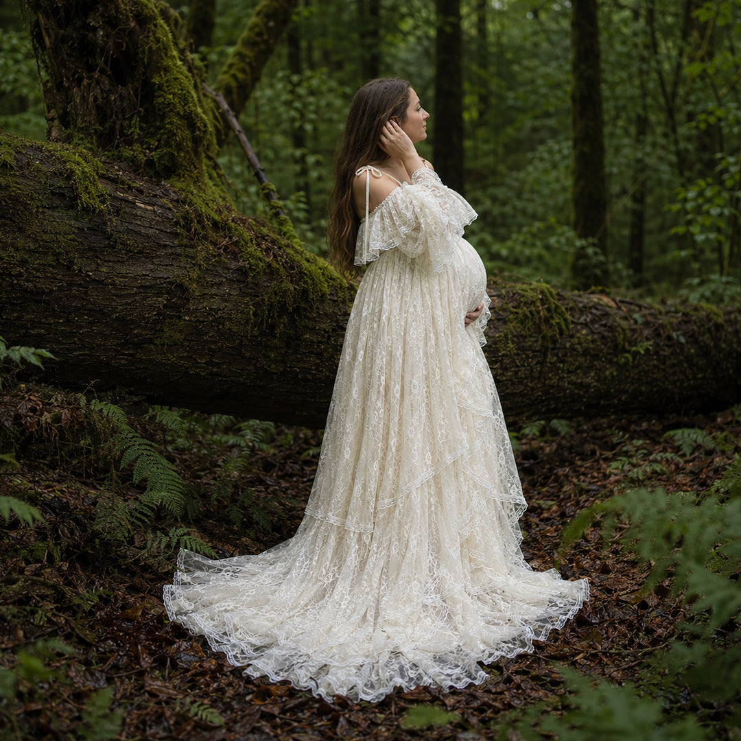 A pregnant woman stands in forest landscape wearing a Melinoe flowing ivory lace maternity gown with off the shoulder ruffled sleeves and a long, tiered skirt that spreads across the leafy ground.