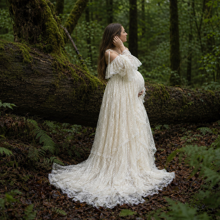 A pregnant woman stands in forest landscape wearing a Melinoe flowing ivory lace maternity gown with off the shoulder ruffled sleeves and a long, tiered skirt that spreads across the leafy ground.