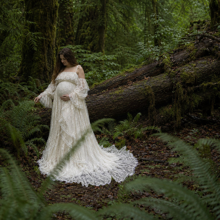 A pregnant woman stands in forest landscape wearing a Melinoe flowing ivory lace maternity gown with off the shoulder ruffled sleeves and a long, tiered skirt that spreads across the leafy ground.