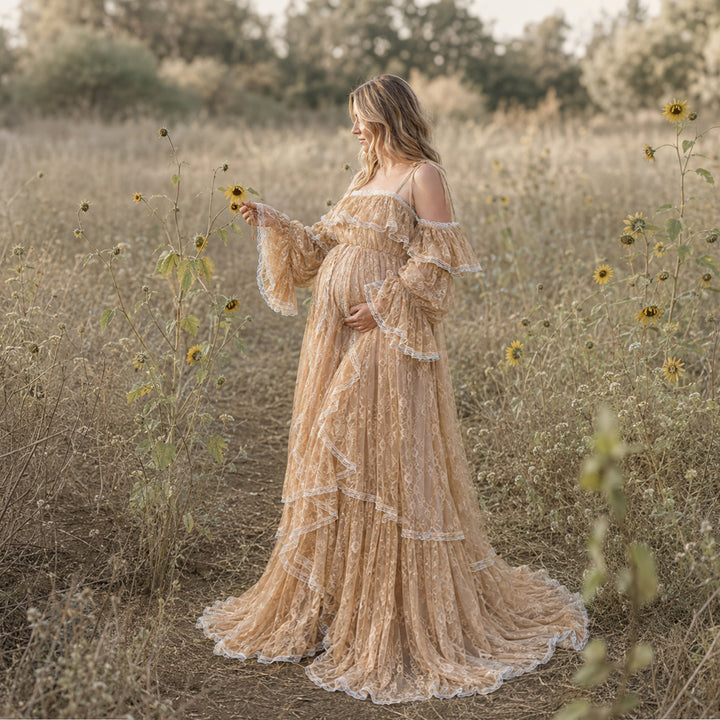 A woman stands in a sunlit field of wildflowers and dry grasses, wearing a long, off the shoulder melinoe gown in a Melinoe soft khaki hue. The dress features delicate lace fabric, tiered ruffle detailing along the bodice and skirt, and flowing bell sleeves, designed specifically as a maternity photography dress.